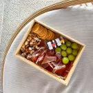 Charcuterie box with Serrano ham, crackers, nuts, dried fruit, grapes, and jam jars.