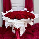 Bouquet of 200 vibrant red roses, elegantly wrapped in white paper, adorned with red ribbons, and set against a backdrop of burgundy fabric.