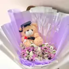 Graduation teddy bear in a bouquet of purple and white dried flowers, wearing a graduation cap.