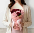 A person holds a pink bouquet with red roses and pink carnations, tied with a pink ribbon.