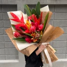 A hand holds a tropical bouquet of red, pink, and yellow exotic flowers, wrapped in brown paper.