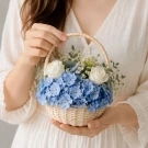 Blue hydrangeas, white roses, and daisies in a woven basket held by hands.