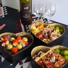 Heart-shaped box overflowing with strawberries, cherries, and chocolates, beside a wine bottle and glasses.