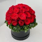Round black box overflowing with 40 vibrant red roses and green foliage.