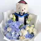 Teddy bear in graduation gown holding a bouquet of blue hydrangeas and white roses.