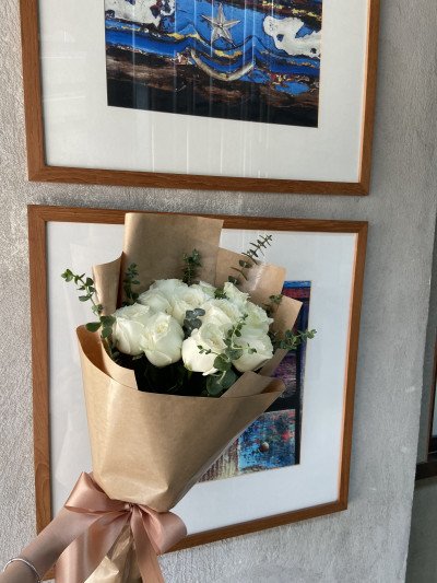 A bouquet of 12 white roses and eucalyptus, wrapped in brown paper with a satin ribbon, held in front of two framed artworks.