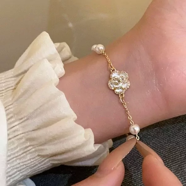 Gold bracelet with a white floral charm and pearls on a wrist.