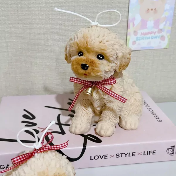 A tan poodle candle with a red ribbon and bell sits on a pink book next to a partial view of another candle.