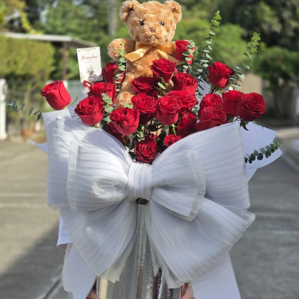 Teddy bear among red roses with a large white bow.