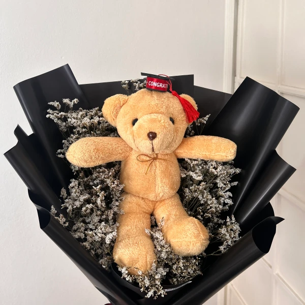 Teddy bear with graduation cap and sash in a bouquet of dried white flowers and black wrapping.