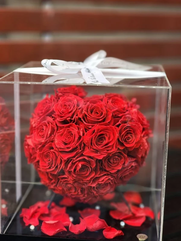 A glass dome encases a heart-shaped arrangement of preserved red roses, tied with a white ribbon.