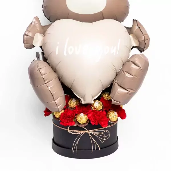 Teddy bear balloon holding heart with "i love you!", surrounded by red carnations and Ferrero Rocher chocolates in a black box.
