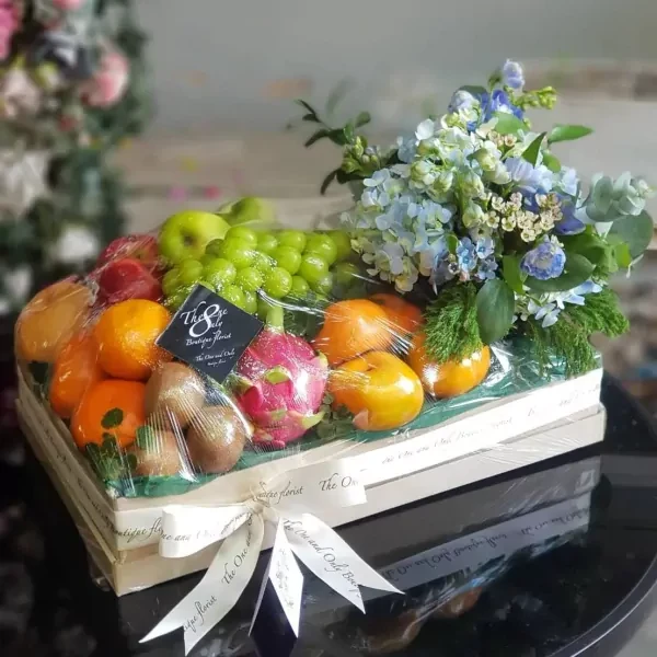 Fruit box with grapes, oranges, dragon fruit, kiwi, and blue flowers, wrapped in plastic.