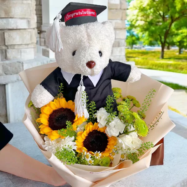 Graduation teddy bear in cap and gown with sunflowers and assorted flowers.