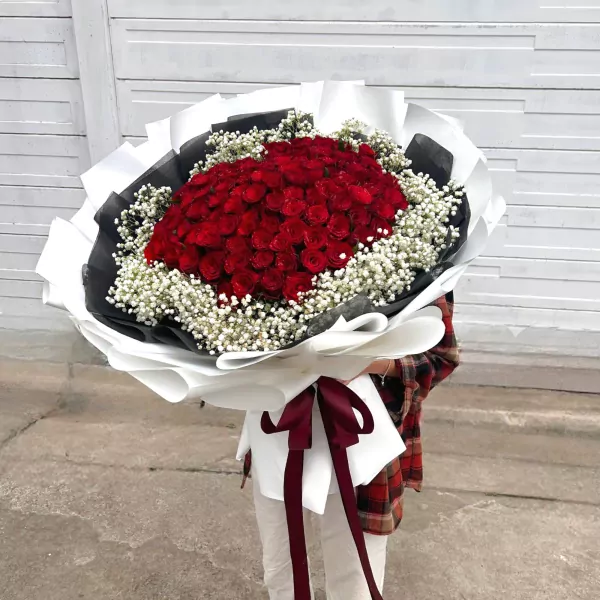 A large bouquet of 99 red roses and baby's breath, wrapped in white and black paper with a burgundy ribbon.