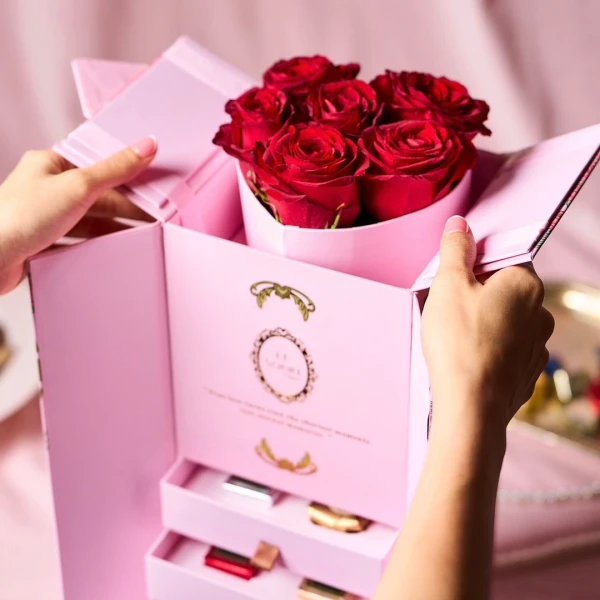 Pink gift box with red roses and chocolate drawers, held by hands.