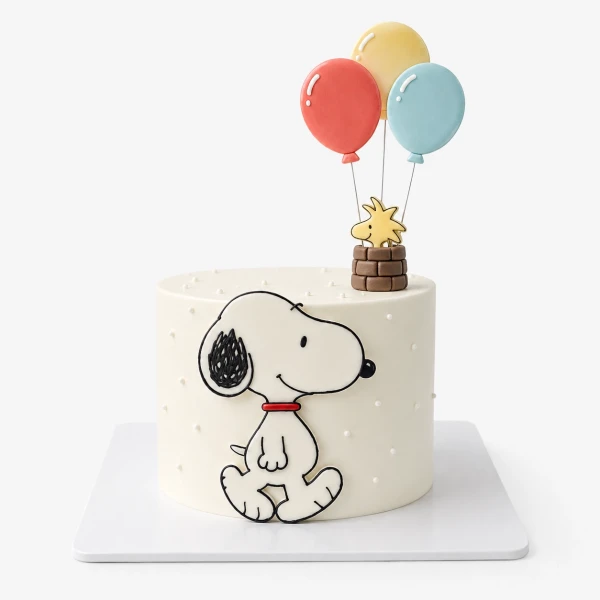 Snoopy and Woodstock float in balloons above a white cake with Snoopy on the side.