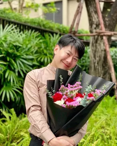 Man in a tan shirt holding a bouquet of red and white roses with purple statice, wrapped in black paper.