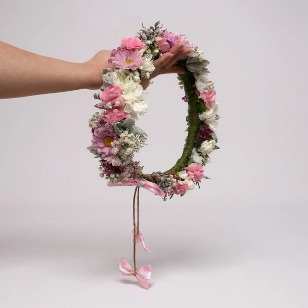 Hand holding a large flower crown with pink and white blooms, featuring a twine ribbon with pink bows.