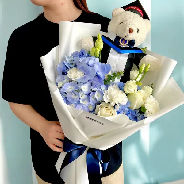 Graduation bouquet with blue hydrangeas, white carnations, and a teddy bear in a graduation gown.