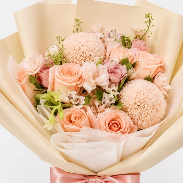 A bouquet of peach roses and pingpong flowers wrapped in ivory paper with a pink ribbon.