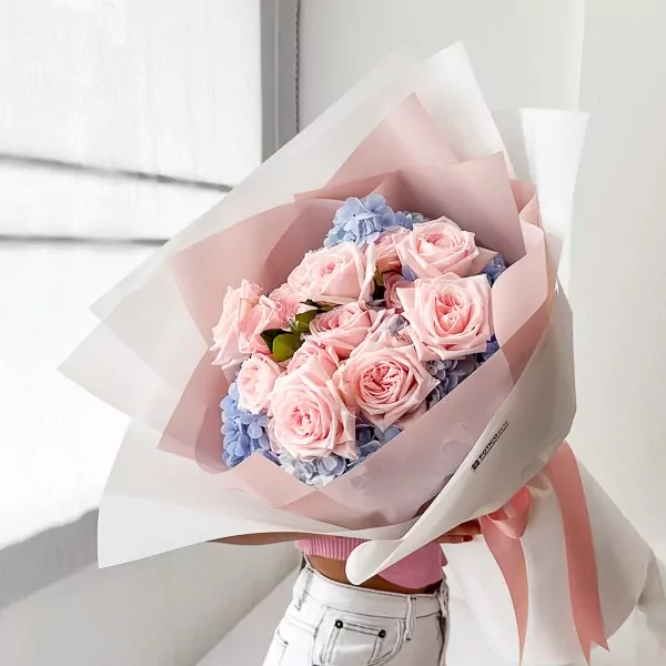 Pink roses and blue hydrangeas in a bouquet with pink and white wrapping paper, tied with a pink ribbon.