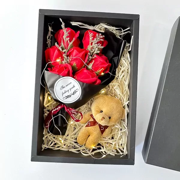 A gift box with red soap roses, a teddy bear keychain, and fairy lights on straw filler.