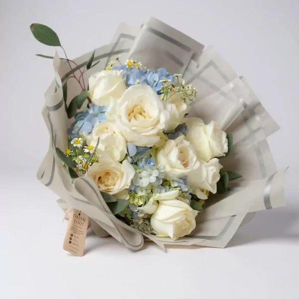 A bouquet of white roses and blue hydrangeas with daisy accents, wrapped in patterned paper with a "Thank You" tag.