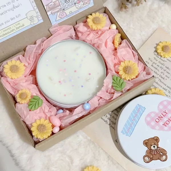 Candle in a tin box surrounded by pink tissue paper and yellow flowers, with a teddy bear tin lid nearby.