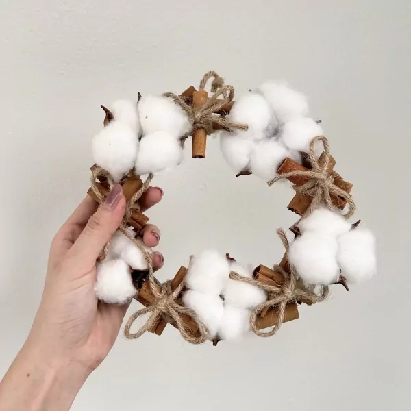 Cotton flower and cinnamon stick wreath with twine bows held by a hand.