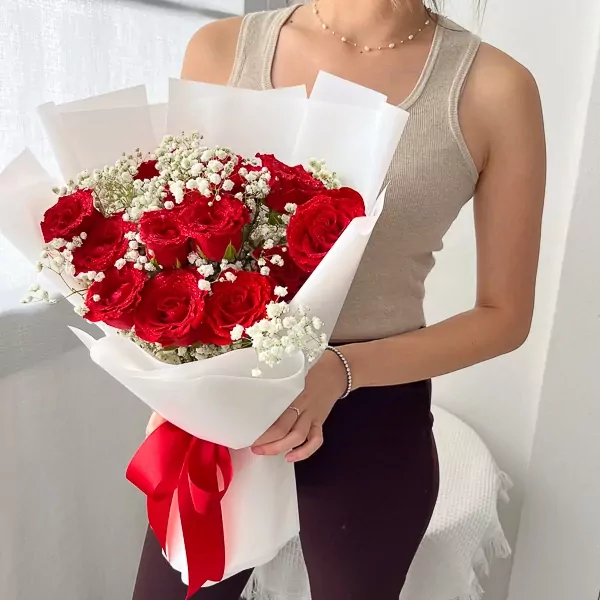 Woman holding a bouquet of 15 red roses with baby's breath, wrapped in white paper and tied with a red ribbon.