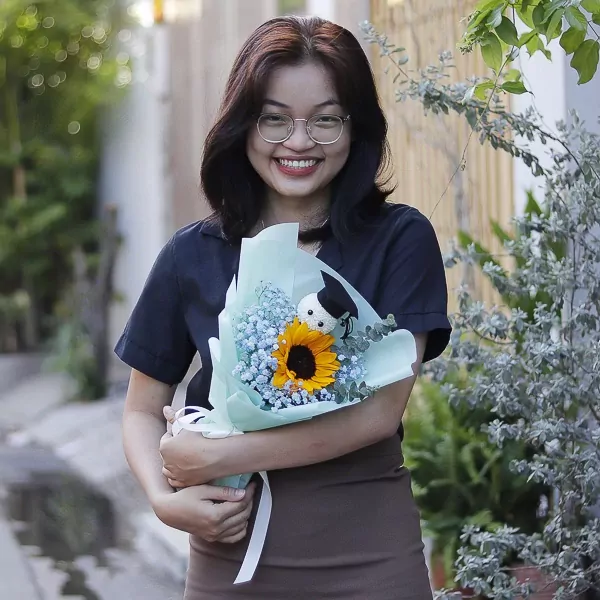 A smiling young woman in glasses holds a graduation bouquet with a sunflower and a small stuffed bear wearing a graduation cap.