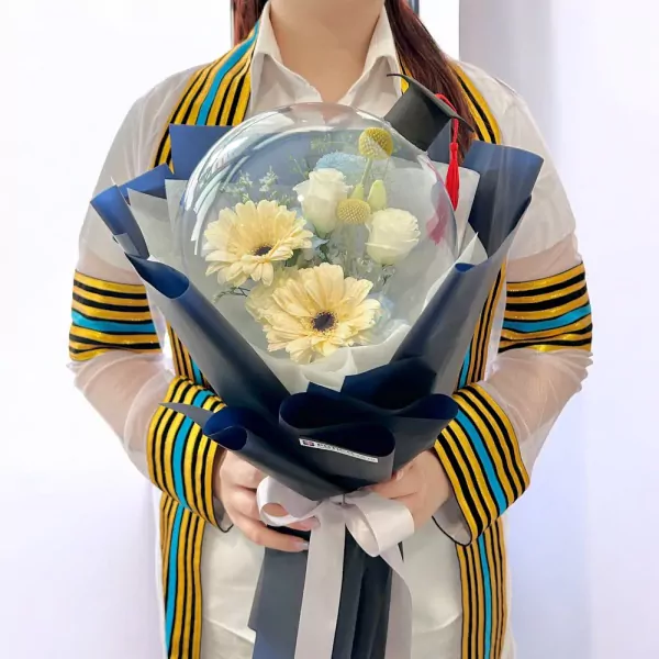 Graduation bouquet in a clear balloon with yellow gerberas and white roses, topped with a mini graduation cap.
