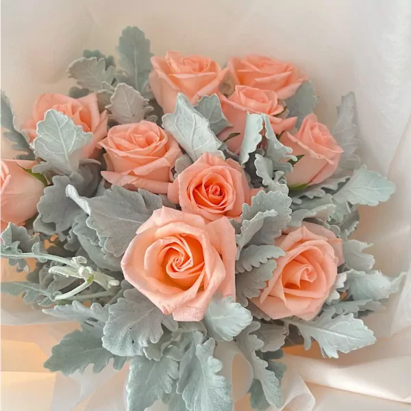 Peach roses with dusty green leaves arranged in a bouquet, wrapped in white paper.