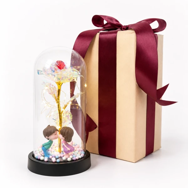 Glass dome with holographic rose, kissing couple figurine, and fairy lights next to a wrapped gift box.