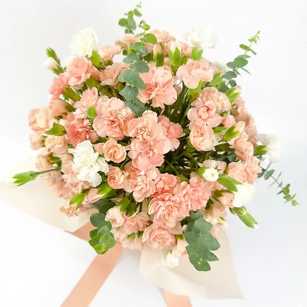 Peach and white carnation bouquet with eucalyptus in a vase, tied with a peach ribbon.