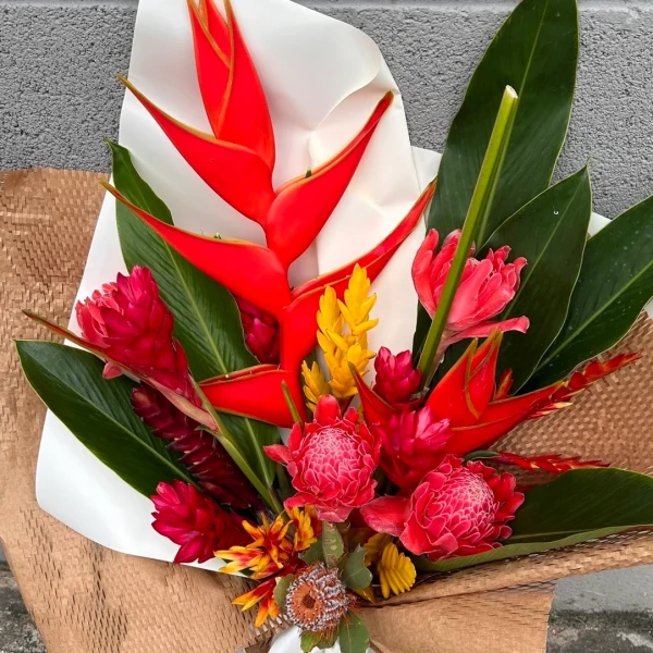 A vibrant bouquet of red and pink tropical flowers, including heliconia, ginger, and torch ginger, with green leaves.