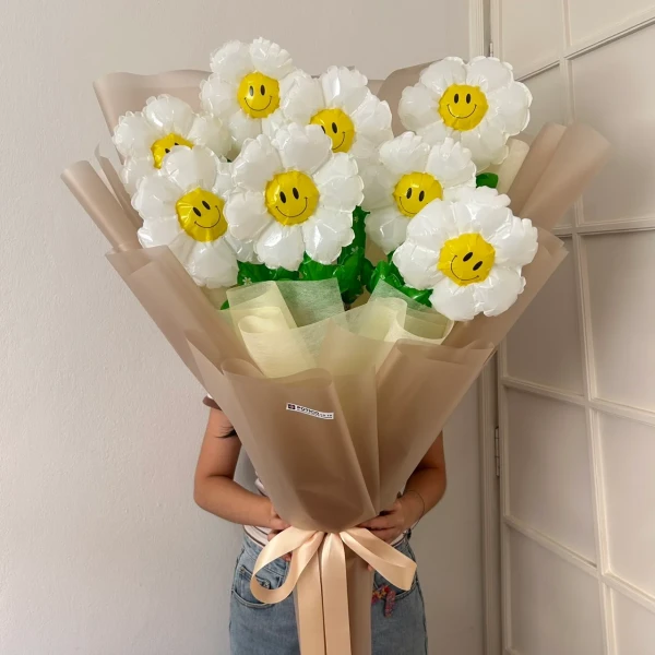 A person holds a large bouquet of white daisy balloons with smiley faces, wrapped in brown paper and tied with a ribbon.