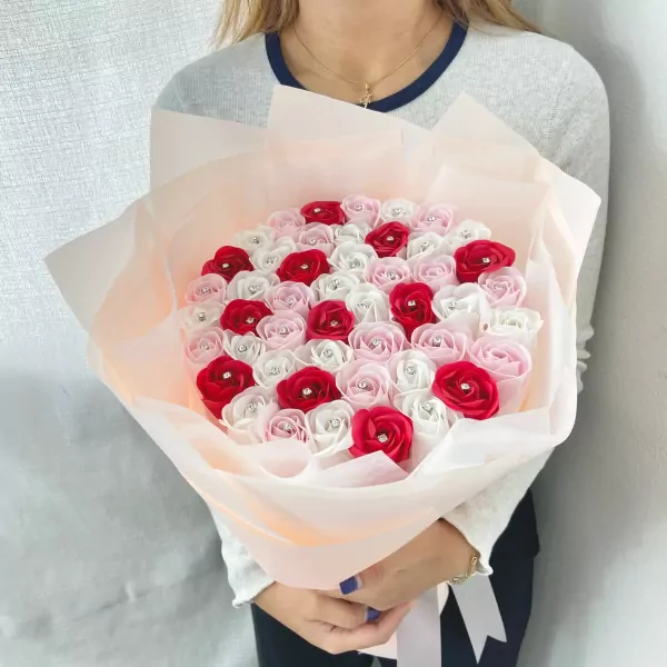 Woman holding a large bouquet of red, pink, and white satin roses with diamond centers, wrapped in pale peach paper.