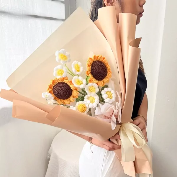 Crocheted sunflower bouquet with white flowers, wrapped in light brown paper, held by a person in a black top and white pants.