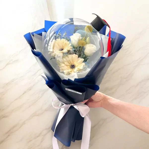 Graduation bouquet in a clear balloon with blue cap, yellow and white flowers, wrapped in dark blue paper.