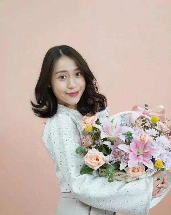 Woman holding a basket of peach and pink lilies, roses, and mums, with a pink background.