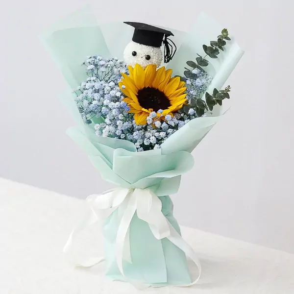 Graduation bouquet with a sunflower, baby's breath, and a graduation cap-wearing plush toy, wrapped in mint paper and tied with a white ribbon.