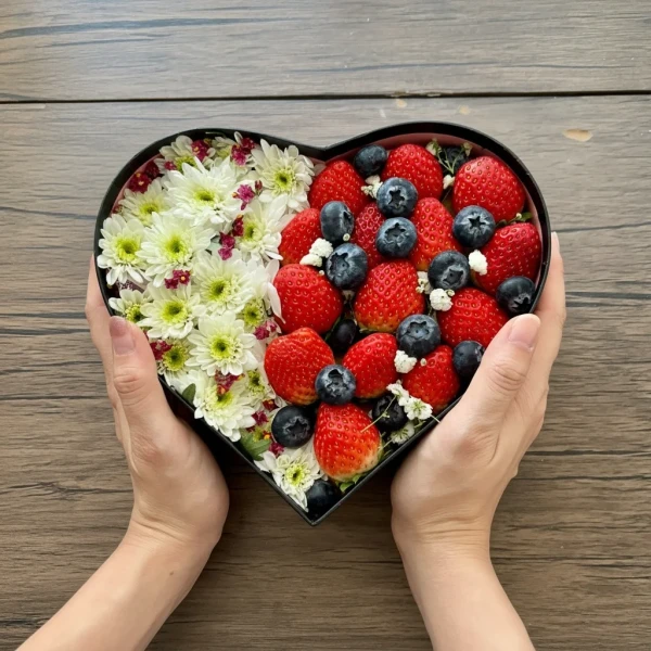Heart-shaped box filled with white flowers, strawberries, and blueberries.