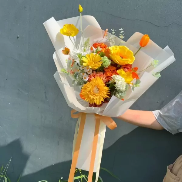 A bright bouquet of yellow and orange artificial flowers wrapped in white paper and tied with a ribbon.