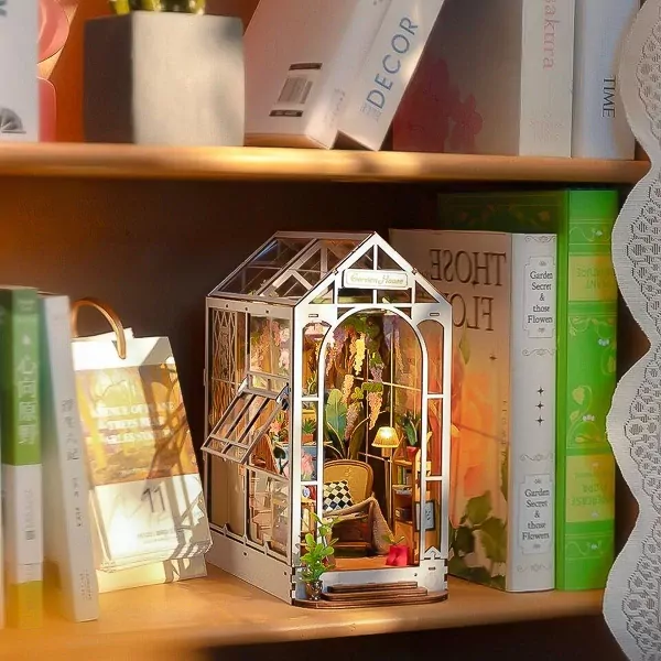 A miniature greenhouse book nook with plants, chair, and lamp, on a shelf with books.