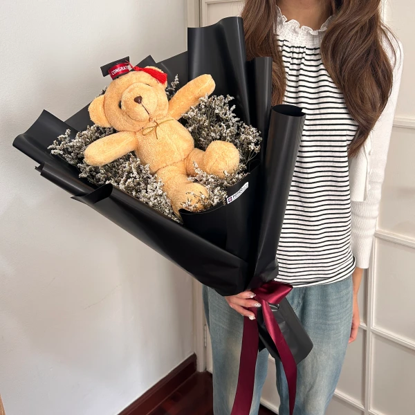 A teddy bear with a graduation hat sits in a bouquet of dried flowers, wrapped in black paper with a burgundy ribbon.