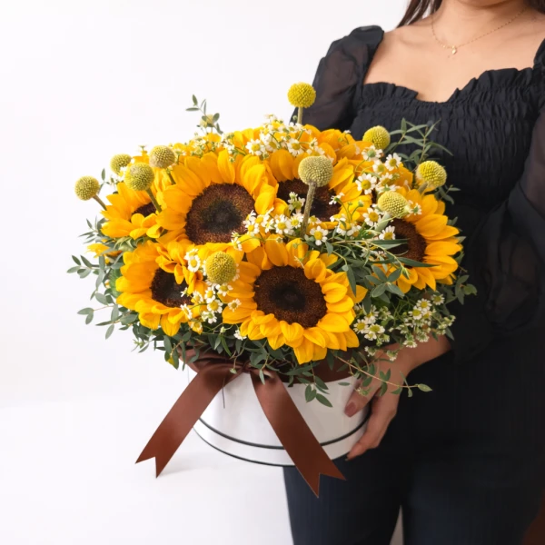 A round white box overflowing with bright yellow sunflowers and small white flowers, tied with a brown ribbon.