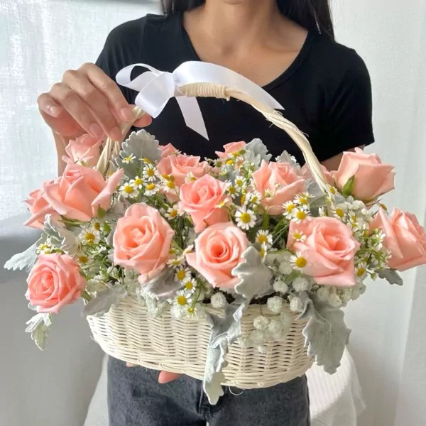 Peach roses and daisies in a woven basket, tied with a white ribbon, held by a person.