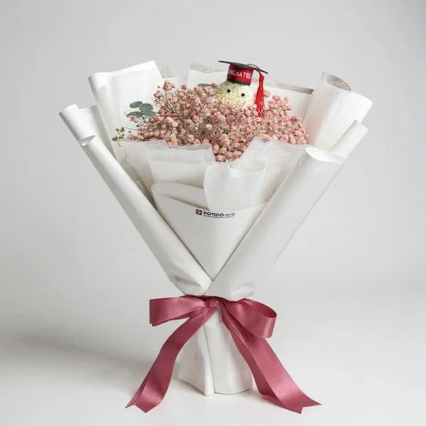 Graduation bouquet with a plush teddy bear wearing a graduation cap, surrounded by delicate pink baby's breath flowers, tied with a pink ribbon.